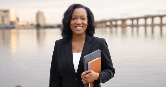 Tavanya holds portfolio and smiles in front of body of water and bridge in the distance
